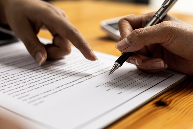 picture is of one persons hand pointing to a signature section of a contract, and another hand of a person with a pen about to sign the contract.