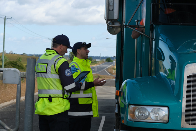 Image is of two Safety Compliance Officers pulled over on the side of the road, standing next to an oversize combination. They are speaking to the driver of the combination