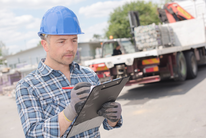 Image is of a man in a hard hat looking at a checklist on a clipboard with a truck in the foreground.